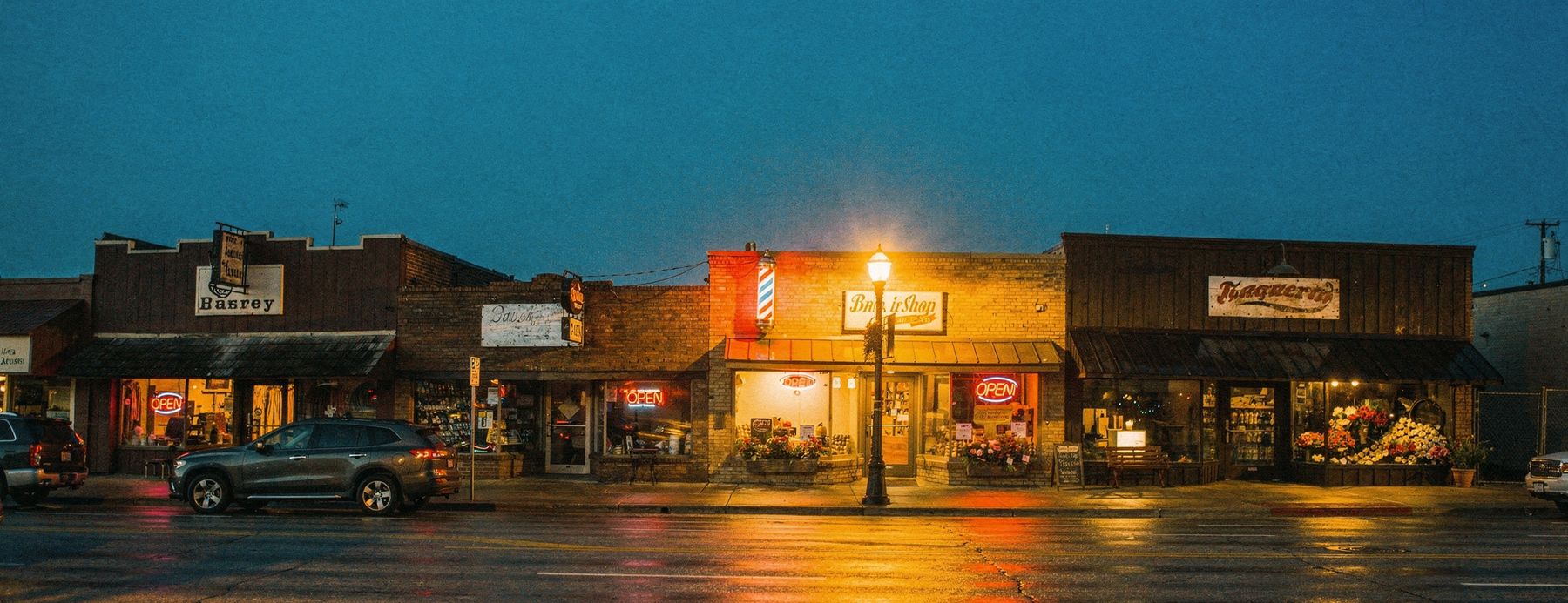 A row of small independent main street shops after dark, windows lit and OPEN signs on